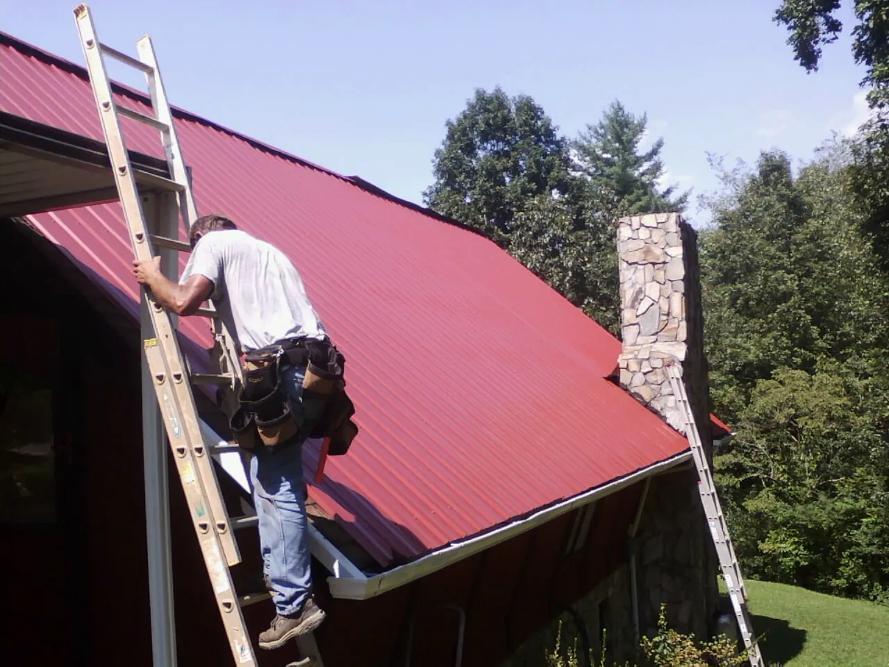 Roofer on a ladder beside a red metal roof and stone chimney — Soffit & Fascia Repair by Roofing Brothers in Marble